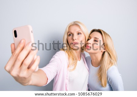 Funky happy cute lovely mother and daughter shooting self portrait on front camera of smart phone sending blowing kiss with pout lips having video-call isolated on grey background