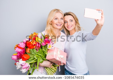 Cheerful joyful blogger having fun with mom using smart phone shooting self postrait on front camera video call enjoying event holding colorful tulips gift case in package isolated on grey background