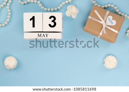 Mother's Day card concept. Wooden calendar and present in craft wrapping with heart and text MOM, ribbon, pearl necklace and white paper flowers on the light blue background. Copy spase, top view