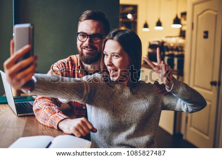 Happy emotional hipster bloggers in casual apparel making funny selfies on mobile application installed on smartphone for publication in social networks.Cheerful male and female students taking photos