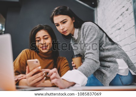 Hipster girl helping her friend installing useful app for learning languages during studying in college, colleagues booking tickets via smartphone online on coffee break together choosing best price Royalty-Free Stock Photo #1085837069