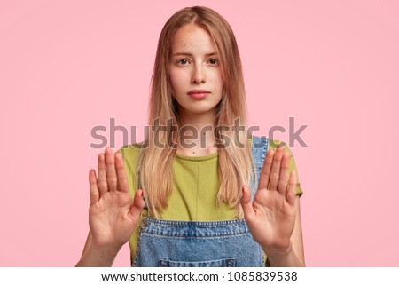 Young attractive female shows prohibition symbol or stop sign, refuses something, shows her refusal or rejection, dressed in jean overalls, poses against pink studio background. Body language concept