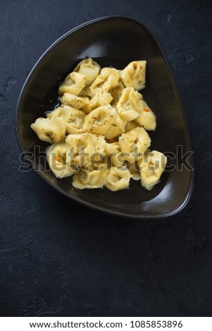 Tortellini in bouillon served in a black bowl, high angle view over black stone background, vertical shot