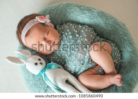 Lovely newborn girl in dress sleeping on a turquoise fluffy background, on a pink flower head. with a knitted white hare toy