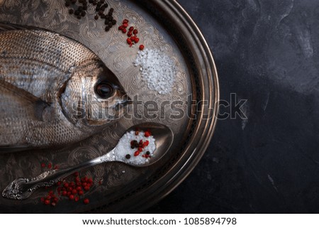 a large fresh sea fish. Fresh fish with a rosemary branch, black pepper, red pepper, salt on a silver platter on a dark background. Beautiful still life picture of food in a low key.