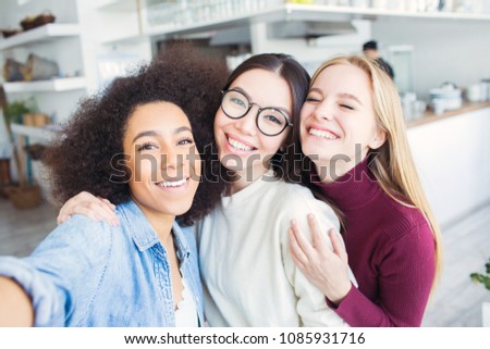 Three beautiful friends are taking selfie together. There are afro american girl, one brunette and one blonde. THey are looking to the camera and smiling. Girls are very cheerful.