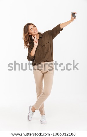 Full length portrait of a smiling young woman taking selfie with mobile phone isolated over white background