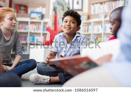 Multiethnic group of kids sitting on floor in circle around the teacher and listening a story. Discussion group of children inaa library talking to woman. Smiling hispanic boy in elementary school. Royalty-Free Stock Photo #1086016097