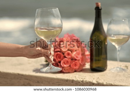 Closeup of female hand green bottle one wedding coral rose bouquet and two glasses with white wine standing on parapet sunny day outdoor on wavy blue ocean or sea water background, horizontal picture