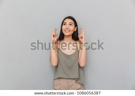 Portrait of a smiling asian woman holding fingers crossed for good luck and looking up isolated over gray background