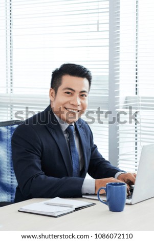 Waist-up portrait of handsome Asian businessman wearing classical suit posing for photography with wide smile while finishing ambitious project on laptop, modern office interior on background