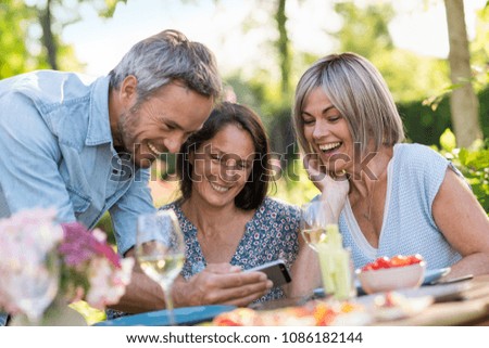 In summer. Group of friends gathered around a table in the garden to share a meal. They have a good time by looking at pictures on a phone