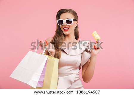 Photo of stylish woman in dress and sunglasses smiling while holding different shopping bags and credit card in hands isolated over pink background
