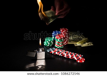 Gambling compostion. Male hand is holding the burning dollar banknote over the iron lighter, money and stacks of poker chips at the black background