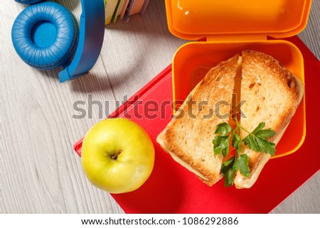 Yellow sandwich box with toasted slices of bread, cheese and green parsley, green apple, headphones and hardback books on the background. School breakfast.