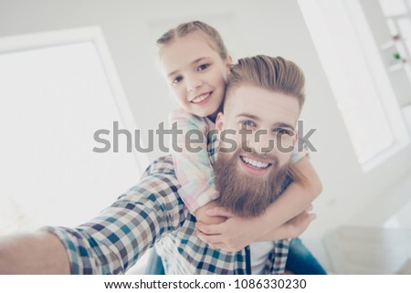 Self portrait of cheerful stylish father with red hair carrying on back cute little schoolgirl, shooting selfie on front camera, hugging embracing, enjoying time together indoor in house, video-call