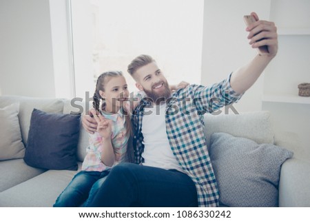Cute girl with bearded stylish father in casual outfit checkered shirts sitting on couch in house embracing shooting self portrait on front camera showing peace symbol holding cell smart phone in hand