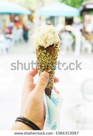 Refreshing ice cream in a chocolate waffle horn in the hand. Hot sunny day on the journey - insta photo for the traveler's blog. Soft focus and beautiful bokeh, tables of cozy cafe in the background.
