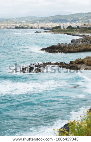 Blue sea waves on a rocky beach, Crete, Greece. Mediterranean coast. Travel and vacation on the beach. Beautiful seascape with small town on the background.