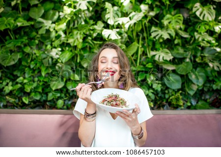 Young and beautiful woman enjoying heathy salad sitting at the vegetarian restaurant with living wall of green plants indoors. Healthy food concept Royalty-Free Stock Photo #1086457103