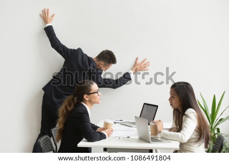 Businessman getting bored of dull work at team meeting, silly office worker leaning on wall pretending funny escape tired of boring routine dreaming of vacation while colleagues having discussion