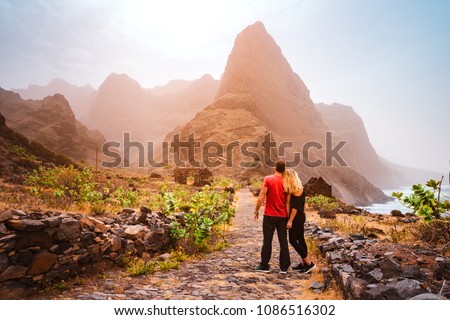 Tourist couple in Aranhas valley hiking from Cruzina to Ponta do Sol. Huge mountains of coastline and old local stone house in the background. Santo Antao Island, Cape Verde Royalty-Free Stock Photo #1086516302