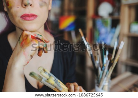 Crop picture of lips and face of beautiful female artist with purple hair and dirty hands with different paints on them, holding paintbrushes in her art studio.