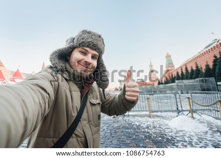tourist man takes a photo of himself against the background of a red square in winter in Moscow, Russia. Shows a thumbs-up, gesture of good humor.