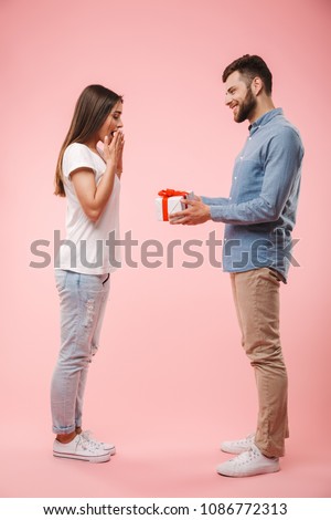 Full length portrait of a cheerful young man giving his girlfriend a present isolated over pink background