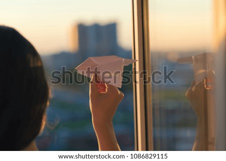 A girl is launching a paper airplane from a window at sunset. 