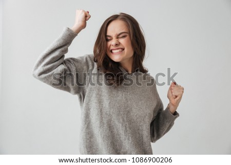 Picture of Pleased brunette woman in sweater rejoices with closed eyes over grey background