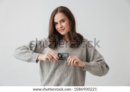 Smiling brunette woman in sweater holding credit card and looking at the camera over grey background