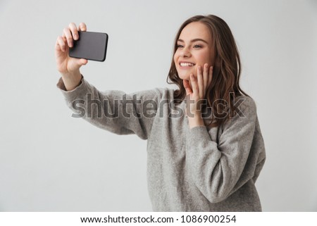 Happy brunette woman in sweater making selfie on smartphone while touching her face over grey background