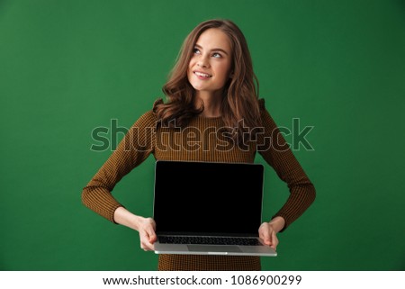 Image of young dreaming woman standing isolated over green background holding laptop computer showing display looking aside.