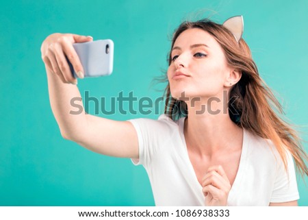 Happy smiling blonde teen girl makes kiss selfie via smartphone on blue background