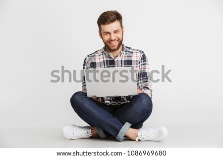 Portrait of a handsome young man in plaid shirt sitting on a floor with laptop computer isolated over white background