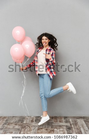 Image of cute beautiful young lady jumping isolated over grey background wall holding balloons pointing.