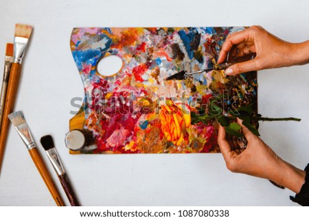 Rose and palette in hands, against the background of brushes and colored pallets. View from above. Artist's work.