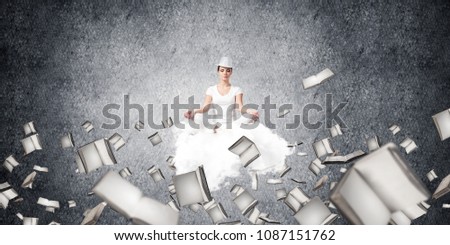 Woman in white clothing keeping eyes closed and looking concentrated while meditating on flying cloud among flying books with gray dark wall on background.