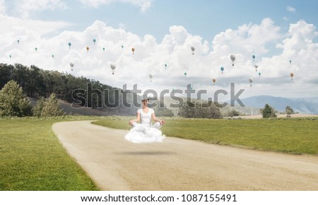 Young woman keeping eyes closed and looking concentrated while meditating on cloud above the road with beautiful and breathtaking landscape on background.