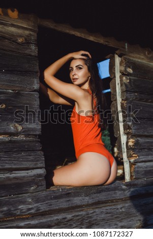 Beautiful slender brunette in a red swimsuit peeking out of the window of a wooden house in the summer at sunset
