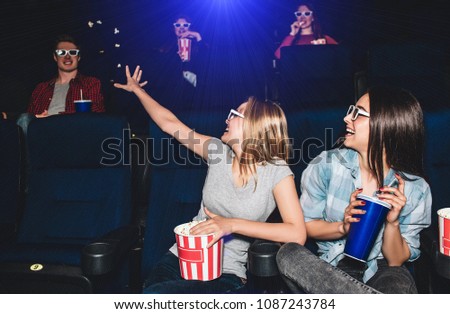 Funny picture of girls sitting together in cinema hall. They are looking at the boy that sits behind them. Blonde girl is trying to reach guy with her hand. Young woman are laughing.