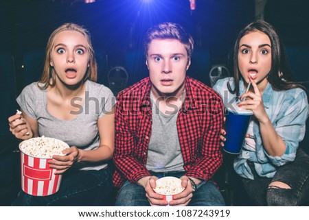 A picture of surprised and amazed young people looking to the camera. Blonde girl is holding a basket of popcorn while brunette girl has a coke in her hands. Man is sitting between them.