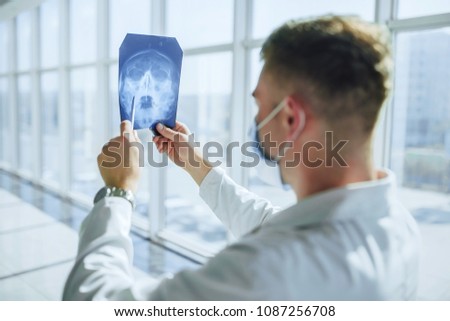Young doctor in a white dressing gown and mask in a modern clinic near the window, examining the x-ray of the head