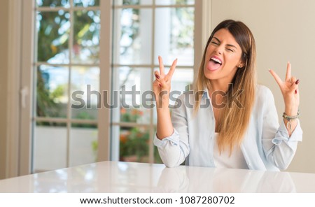 Young beautiful woman at home looking at camera showing tong and making victory sign with fingers
