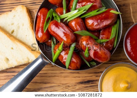 Juicy meat sausages with crispy toast. With green onions, mustard and sauce. In a small saucepan on a wooden board. View from above. Close-up