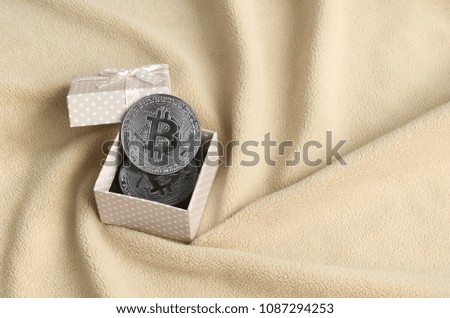 The silver bitcoin lies in a small orange gift box with a small bow on a blanket made of soft and fluffy light orange fleece fabric with a large number of relief folds