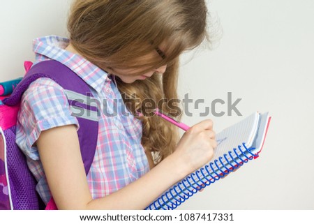 Girl child elementary school student wearing glasses with a backpack writing in her notebook. Bright school wall background