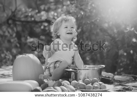 One laughing boy at picnic sitting with ladle pot orange pumpkin red tomato squash and cucumber playing with food sitting on blue checkered plaid on natural background sunny day, horizontal picture