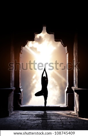 Yoga vrikshasana tree pose by man silhouette in old temple arch at dramatic sunset sky background. Free space for text